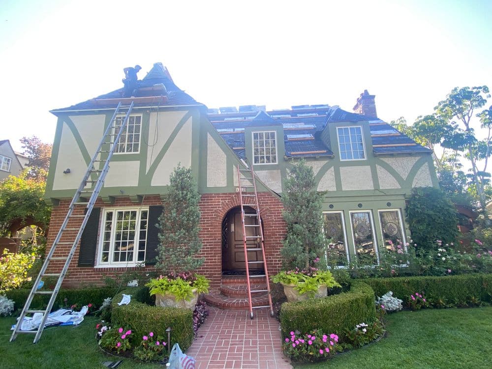 Tudor-style home with roof renovation, ladders, and colorful garden in foreground.