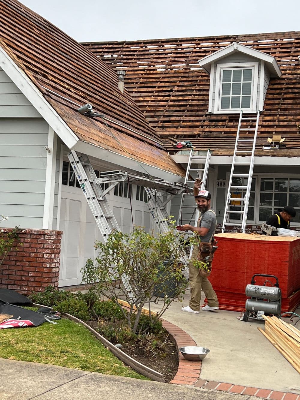 Roofing workers replacing shingles on a residential house with ladders and tools visible.