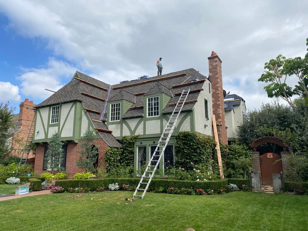 Roof renovation in progress on a Tudor-style house with a worker on the roof and a ladder.