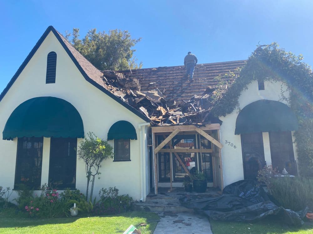Roof repair in progress on a charming white house with green awnings and a sunny sky.