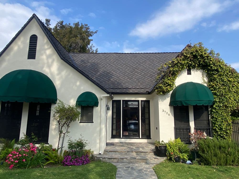 Charming white house with green awnings, stone walkway, and lush garden, sunny blue sky.